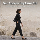 Frau in schwarzer Hose, schwarzem Rollkragenpullover und Ballerinas auf einer Altstadtstraße im Stil von Audrey Hepburn