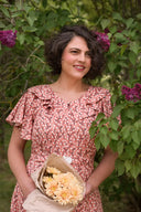 Frau mit kurzen lockigen dunklen Haaren trägt ein rosafarbenes, floral gemustertes Kleid von Emmy Design und hält einen Strauß heller Blumen in braunem Papier, stehend vor grünen Blättern und violetten Blüten im Garten.