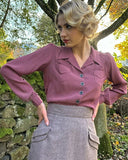 Frau trägt roséfarbene Bluse und grauen Rock von The House of Foxy, steht im Freien vor Steinmauer und herbstlichen Bäumen