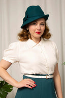 Frau in cremefarbener Vintage-Bluse mit Kragen und kurzen Puffärmeln, kombiniert mit petrolfarbenem Rock, weißem Gürtel und passendem Filzhut